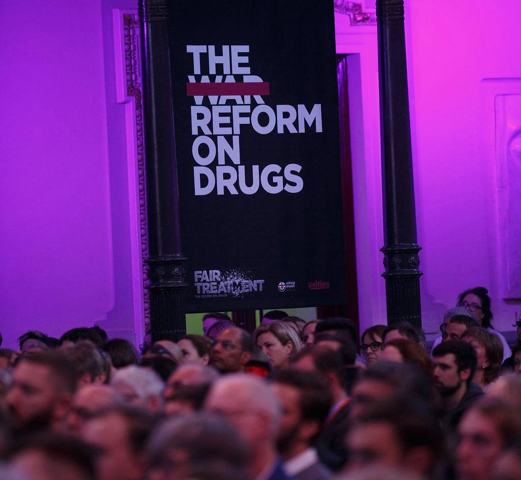 A crowd of diverse people sit in a theatre under a sign that reads 'the reform on drugs'.
