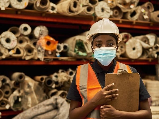 A young worker with brown skin wearing a high-vis vest and holding a clip board.