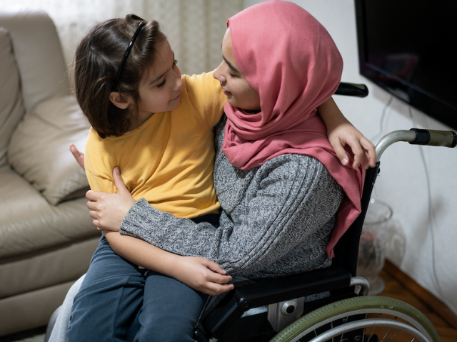 A woman who uses a wheelchair and wears a pink head scarf holding a young child. They are smiling at each other and talking.