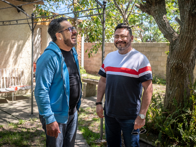 Two middle-aged men standing in a garden together. One man is smiling at the camera; the other is laughing. 