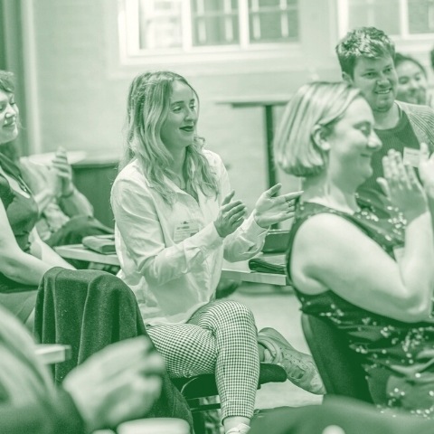 Three younger people sit in chairs in a conference room laughing and clapping.