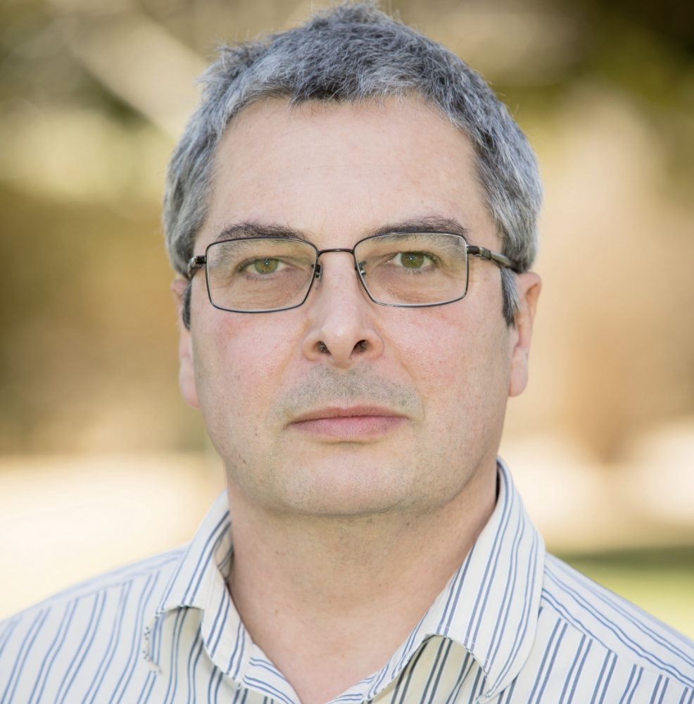 A man with light skin looks at the camera. He wears glasses and has short grey hair.