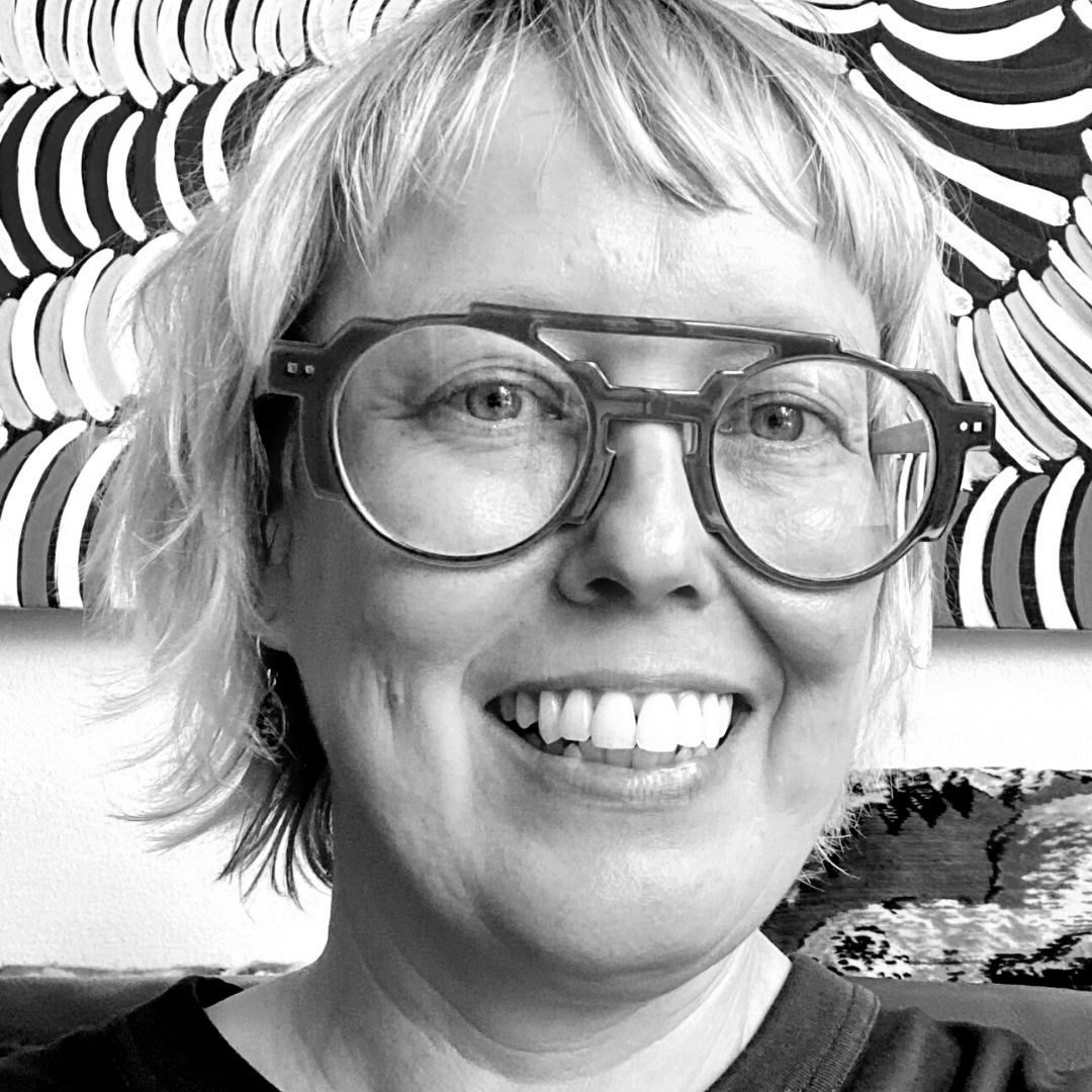 Headshot of a person wearing thick glasses smiling in front of an abstract painting.