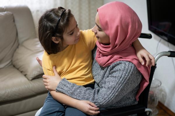 A woman and child smiling at each other. The woman is using a wheelchair and wears a pink headscarf; the child has short dark hair and is wearing a yellow t-shirt.