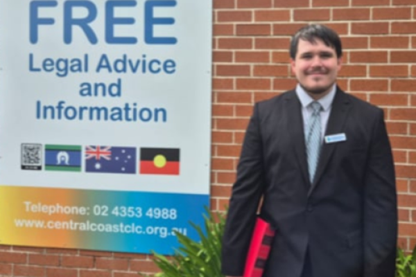 Ivan standing outside the Central Coast CLC building with a sign reading 'Free legal advice and information'. He is smiling and holding a binder.